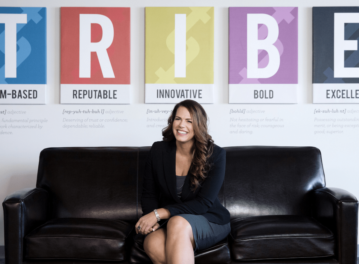 Renee Merrifield smiling and sitting on black couch with T-R-I-B-E posters behind her