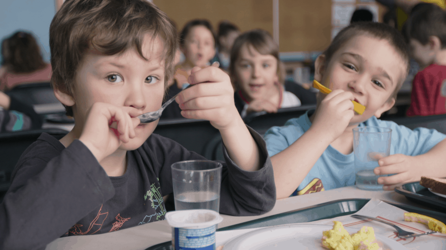 Two kids eating and drinking water with blurred background of other children